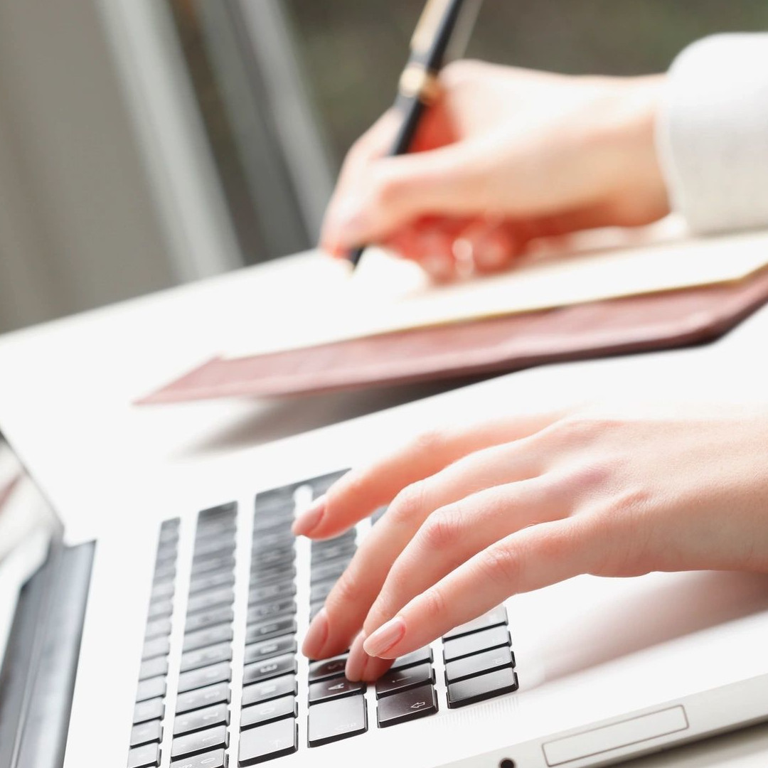 A person typing on a laptop with a pen and paper.